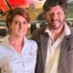 Mila Seidl e Lito Sousa sorrindo juntos, com avião militar ao fundo