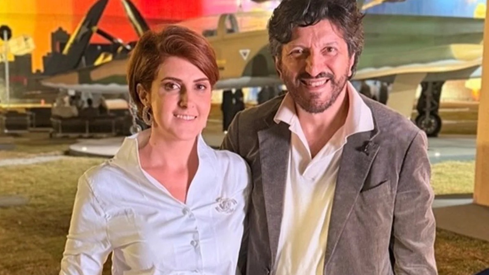 Mila Seidl e Lito Sousa sorrindo juntos, com avião militar ao fundo