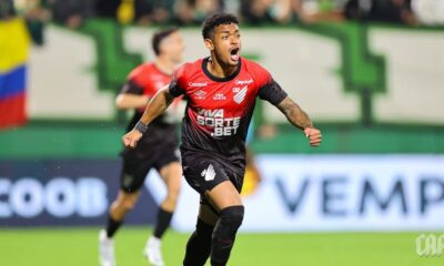 Jogador do Athletico-PR em camisa preta/vermelha (Umbro, Viva Sorte) celebra em campo verde