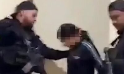Dois policiais em uniforme escuro detêm uma mulher com o rosto borrado e capuz