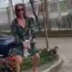 Mulher de óculos escuros e vestido estampado segura um cachorro pequeno na rua sendo filmada
