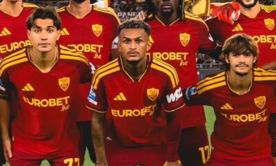 Três jogadores com uniformes vinho e dourado da Roma com logo Adidas e 'EUROBET Live'