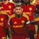Três jogadores com uniformes vinho e dourado da Roma com logo Adidas e 'EUROBET Live'