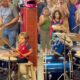 Menino de cabelos encaracolados toca bateria azul profissional em apresentação, público assiste