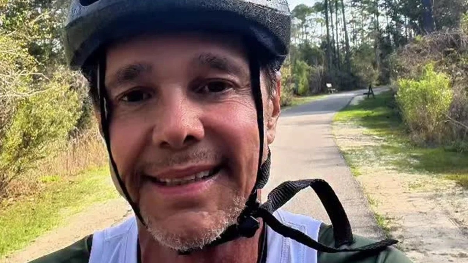 Close-up de homem sorrindo com capacete de bicicleta em trilha externa com árvores