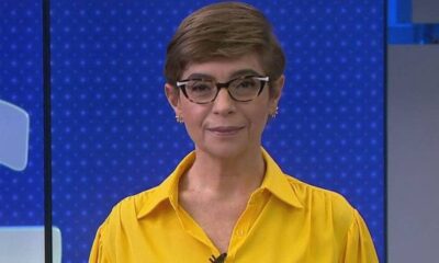 Renata Lo Prete com cabelo curto, óculos e camisa amarela, sorrindo em estúdio de TV