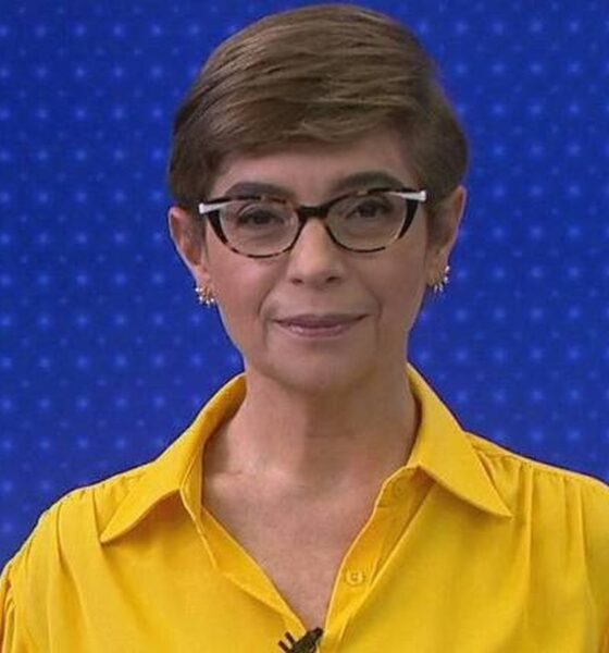 Renata Lo Prete com cabelo curto, óculos e camisa amarela, sorrindo em estúdio de TV
