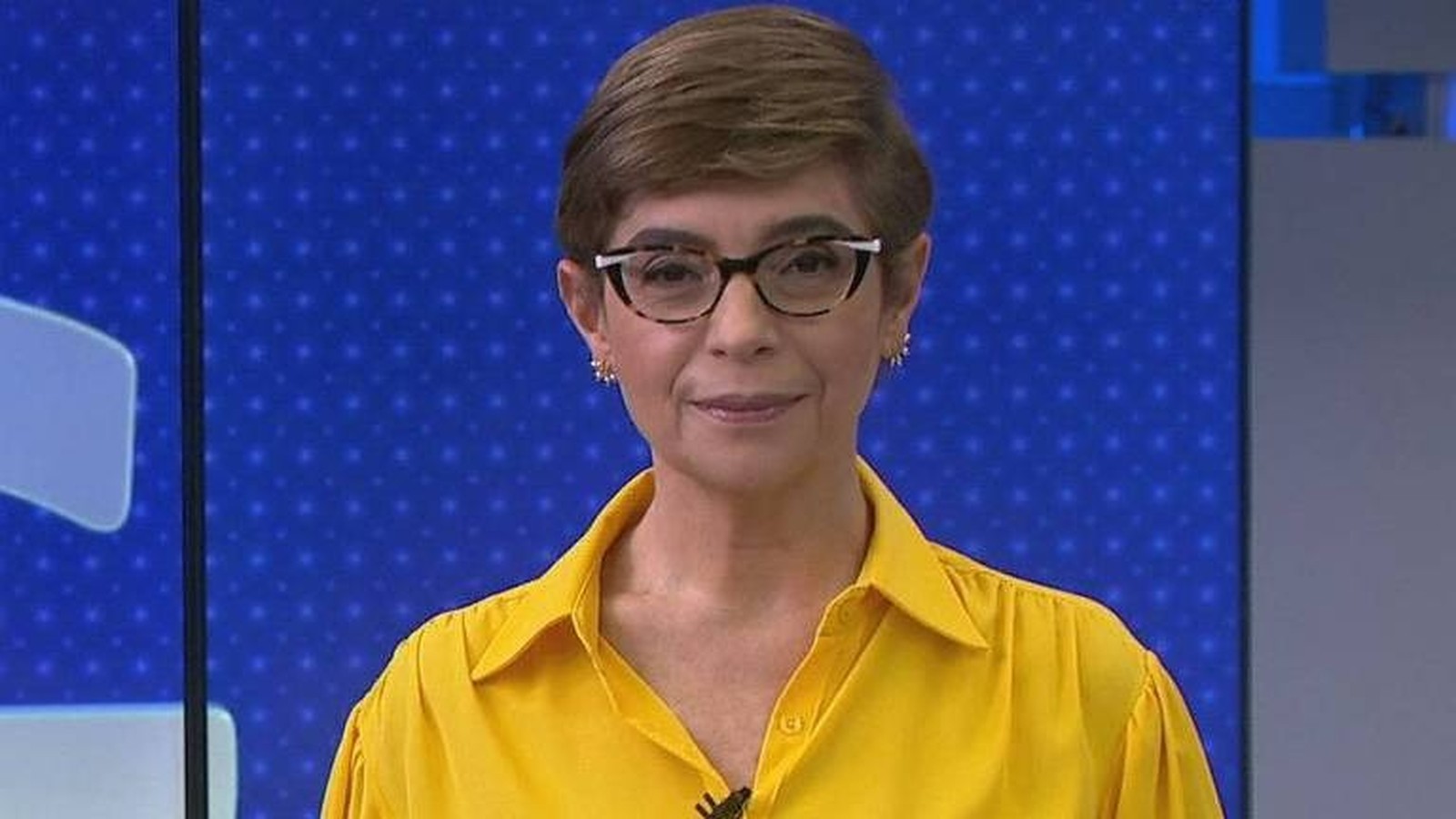 Renata Lo Prete com cabelo curto, óculos e camisa amarela, sorrindo em estúdio de TV