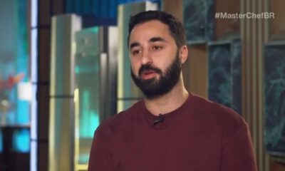 Homem barbudo com camiseta vinho e microfone. Ao fundo, hashtag #MasterChefBR