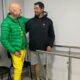 Dois homens sorrindo, um com prótese na perna, ao lado de barras paralelas de reabilitação