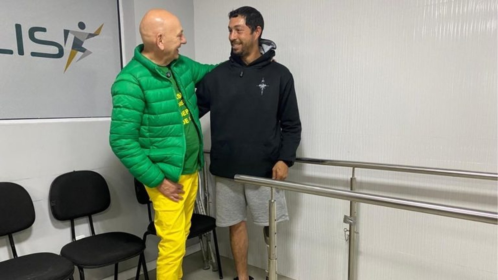 Dois homens sorrindo, um com prótese na perna, ao lado de barras paralelas de reabilitação