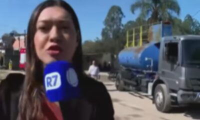 Repórter feminina com microfone azul da R7 em mãos, caminhão tanque azul ao fundo