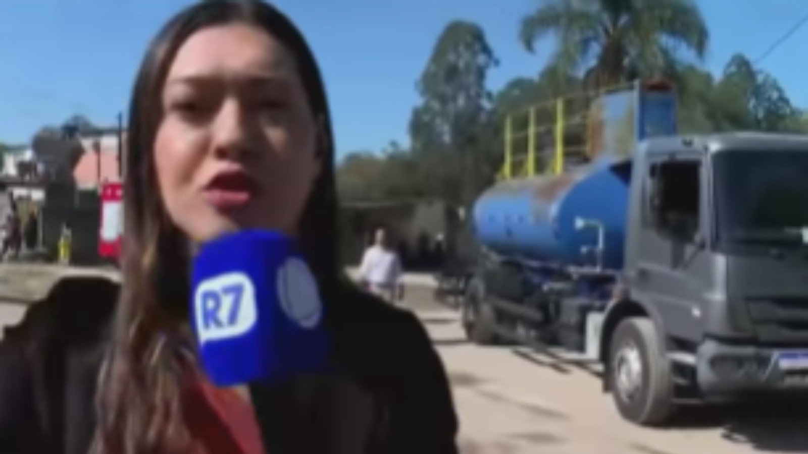 Repórter feminina com microfone azul da R7 em mãos, caminhão tanque azul ao fundo