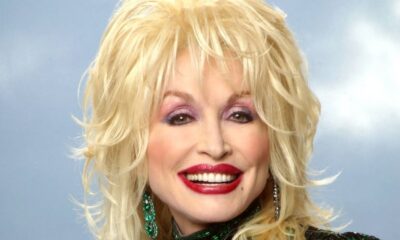 Dolly Parton sorrindo, cabelo loiro, batom vermelho, maquiagem roxa, brincos verdes