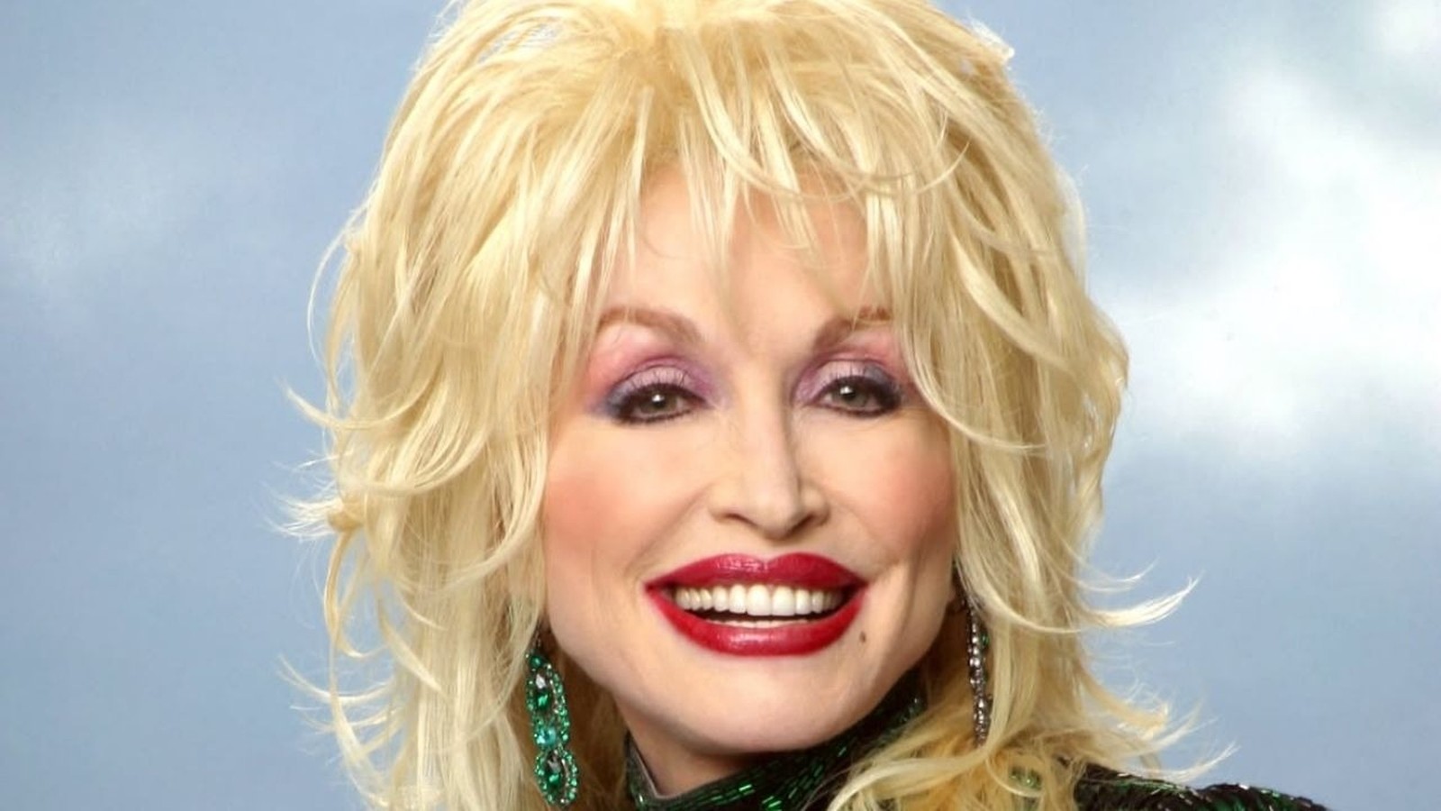 Dolly Parton sorrindo, cabelo loiro, batom vermelho, maquiagem roxa, brincos verdes