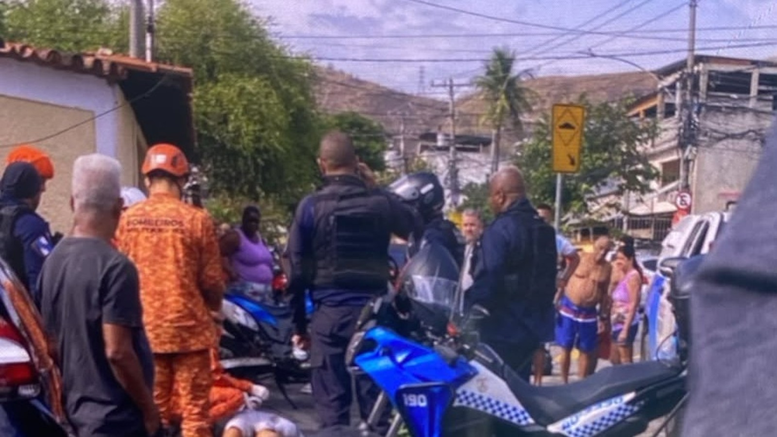 Homens da polícia e bombeiros em uma rua, com motocicletas e um corpo no chão