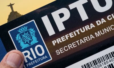 Polegar segura documento IPTU Rio Prefeitura com logotipo municipal e Cristo Redentor desfocado ao fundo