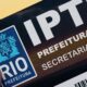 Polegar segura documento IPTU Rio Prefeitura com logotipo municipal e Cristo Redentor desfocado ao fundo