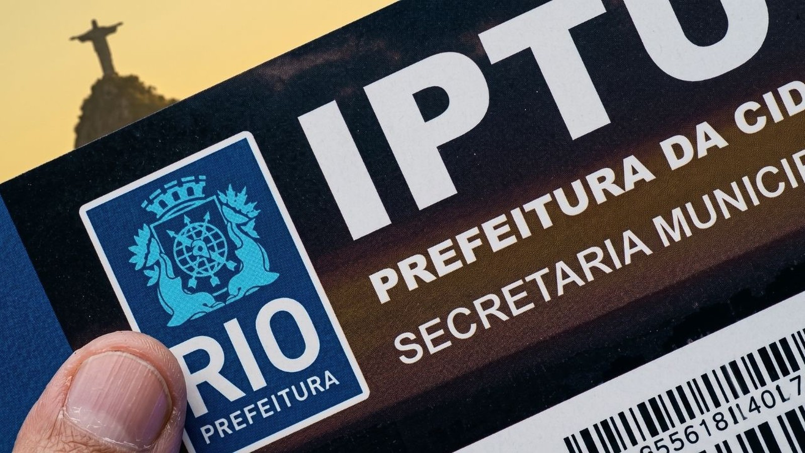 Polegar segura documento IPTU Rio Prefeitura com logotipo municipal e Cristo Redentor desfocado ao fundo