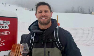 Homem sorridente com jaqueta, mochila e taça de vinho tinto em pista de esqui nevada