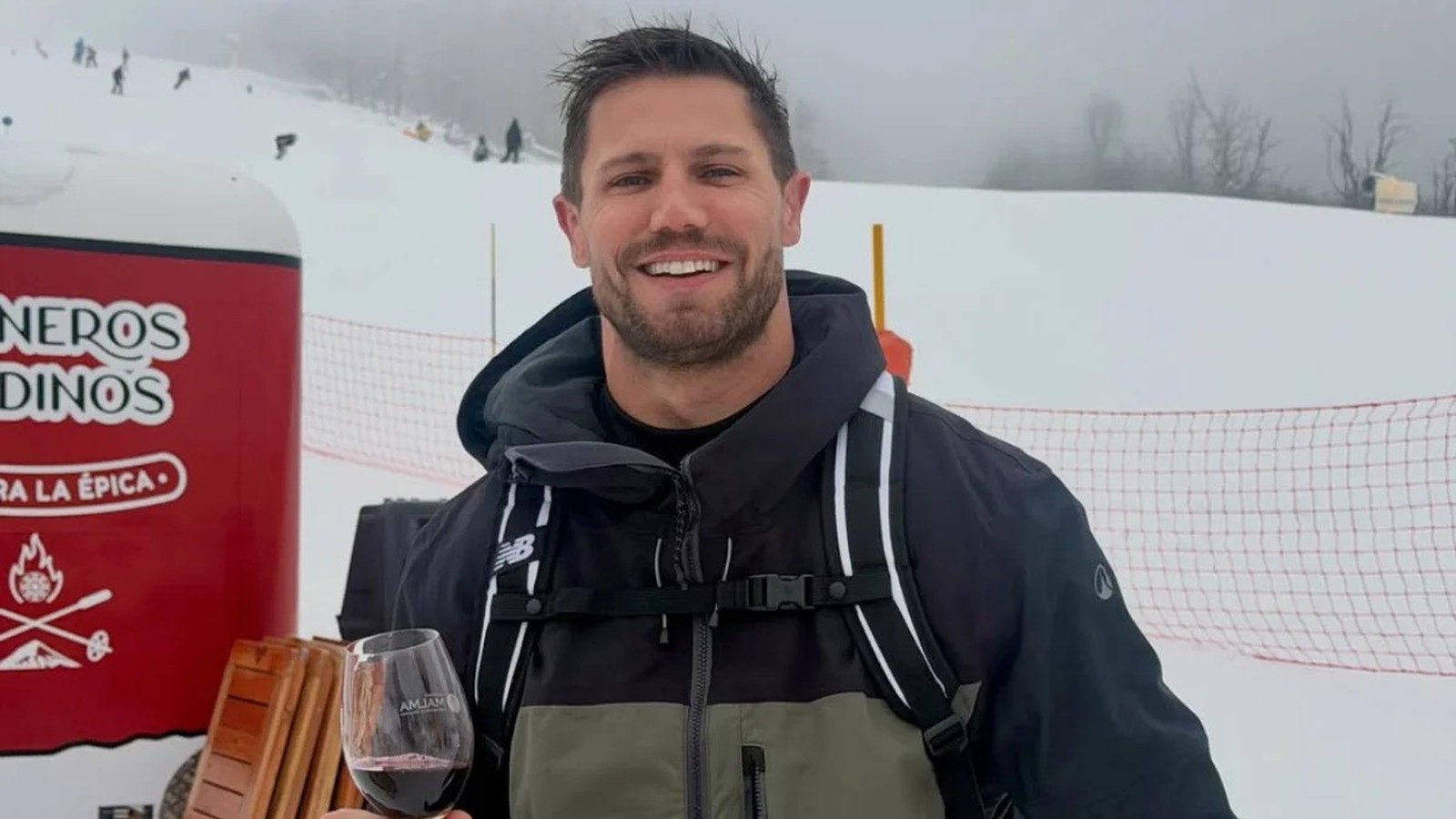 Homem sorridente com jaqueta, mochila e taça de vinho tinto em pista de esqui nevada