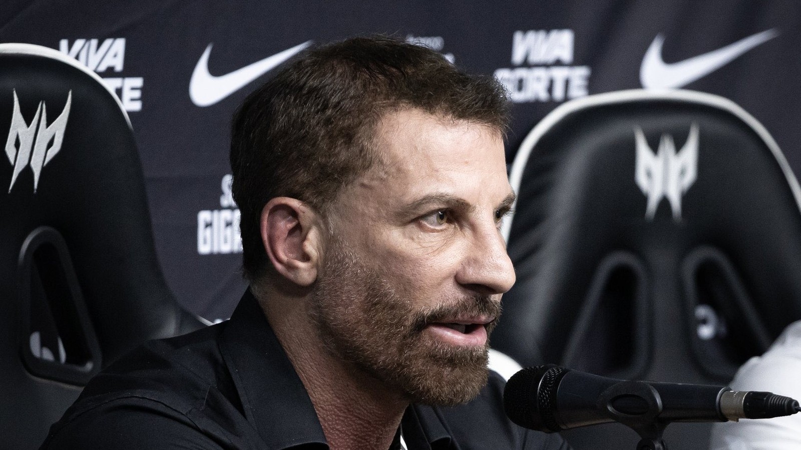Homem barbudo de camisa preta fala ao microfone, fundo com logos Nike e Predator