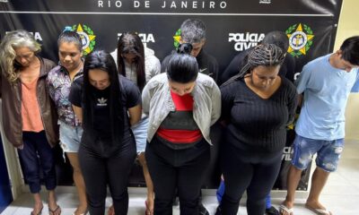 Dez pessoas detidas, maioria mulheres, cabeças baixas, contra banner da Polícia Civil