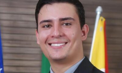 Retrato de homem jovem sorrindo, de terno e camisa, com bandeiras desfocadas ao fundo