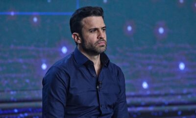 Homem de cabelo e barba escura, camisa azul-marinho, com expressão séria em estúdio
