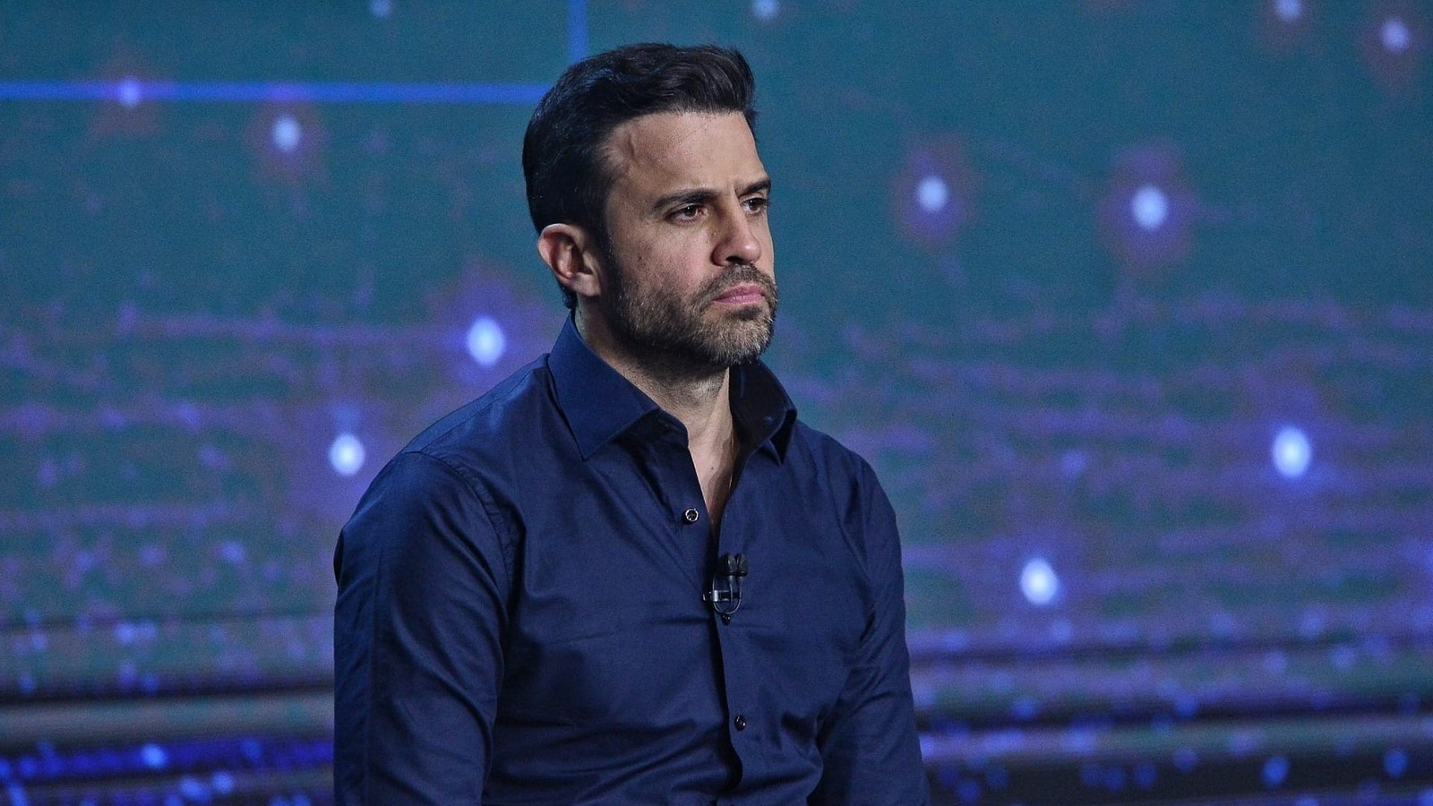 Homem de cabelo e barba escura, camisa azul-marinho, com expressão séria em estúdio