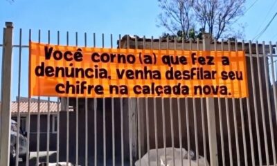 Banner laranja em portão de metal com texto ofensivo sobre denúncia de calçada
