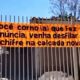 Banner laranja em portão de metal com texto ofensivo sobre denúncia de calçada