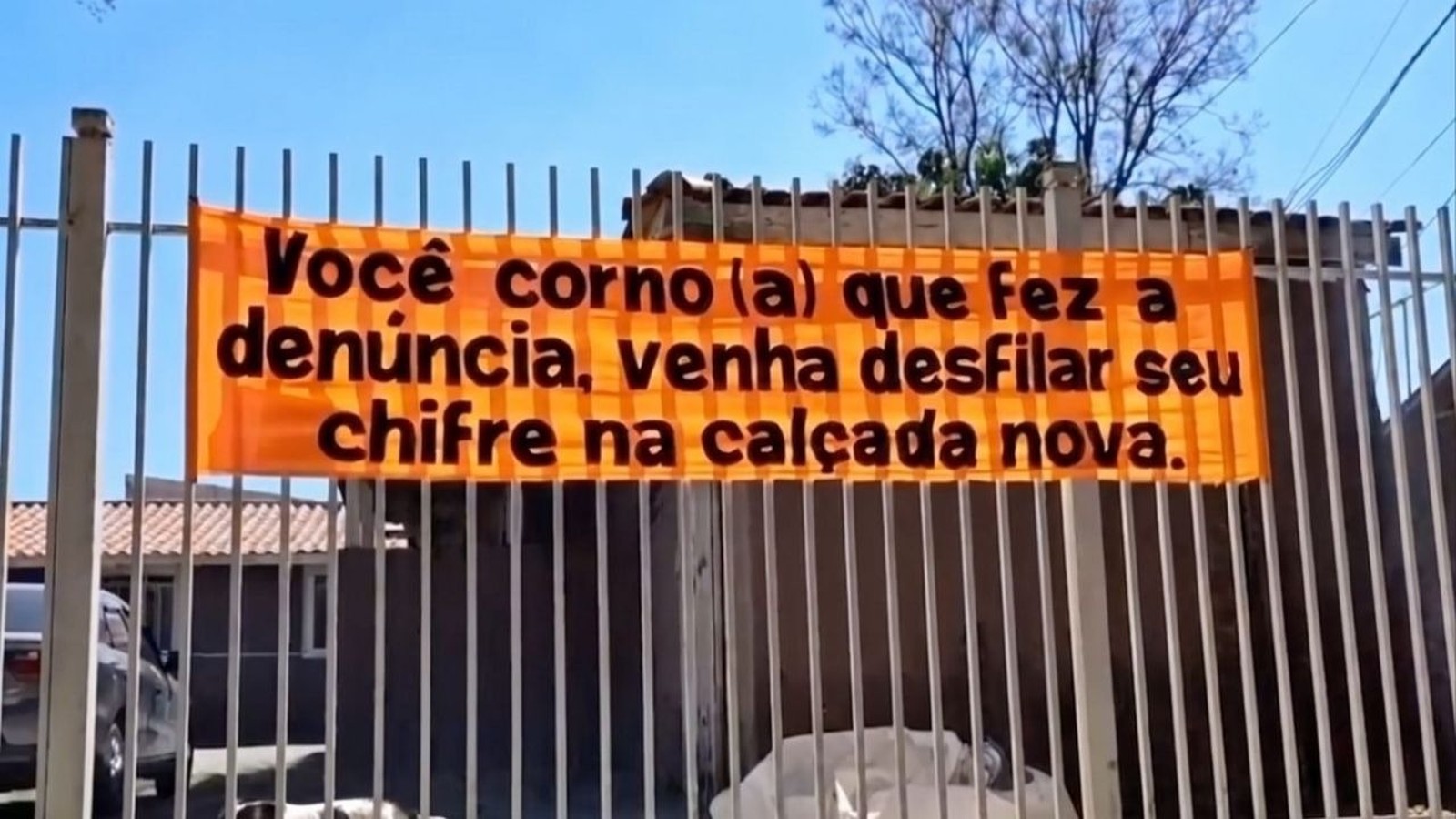 Banner laranja em portão de metal com texto ofensivo sobre denúncia de calçada