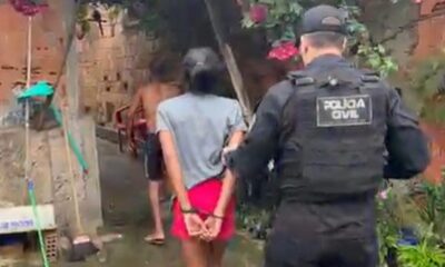 Oficial da Polícia Civil escolta dois detidos, um algemado, em viela com paredes de tijolo