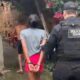 Oficial da Polícia Civil escolta dois detidos, um algemado, em viela com paredes de tijolo