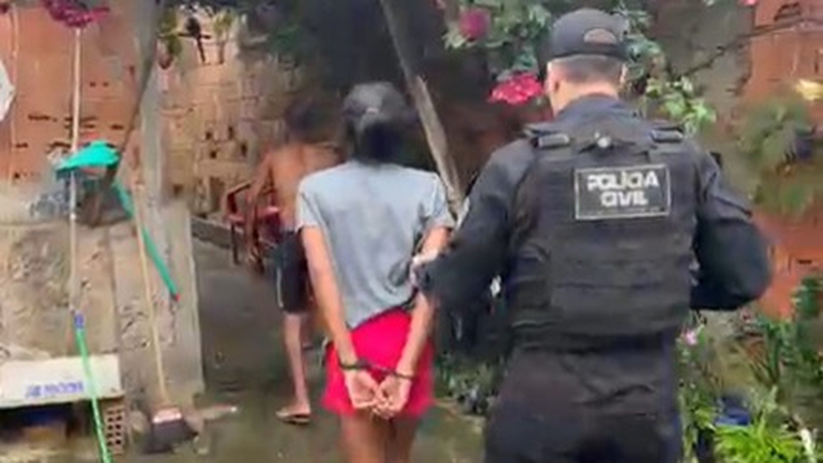 Oficial da Polícia Civil escolta dois detidos, um algemado, em viela com paredes de tijolo