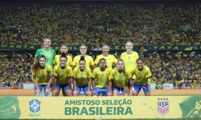 Seleção brasileira feminina posa em campo, estádio lotado. Faixa com "AMISTOSO SELEÇÃO BRASILEIRA", CBF e USA