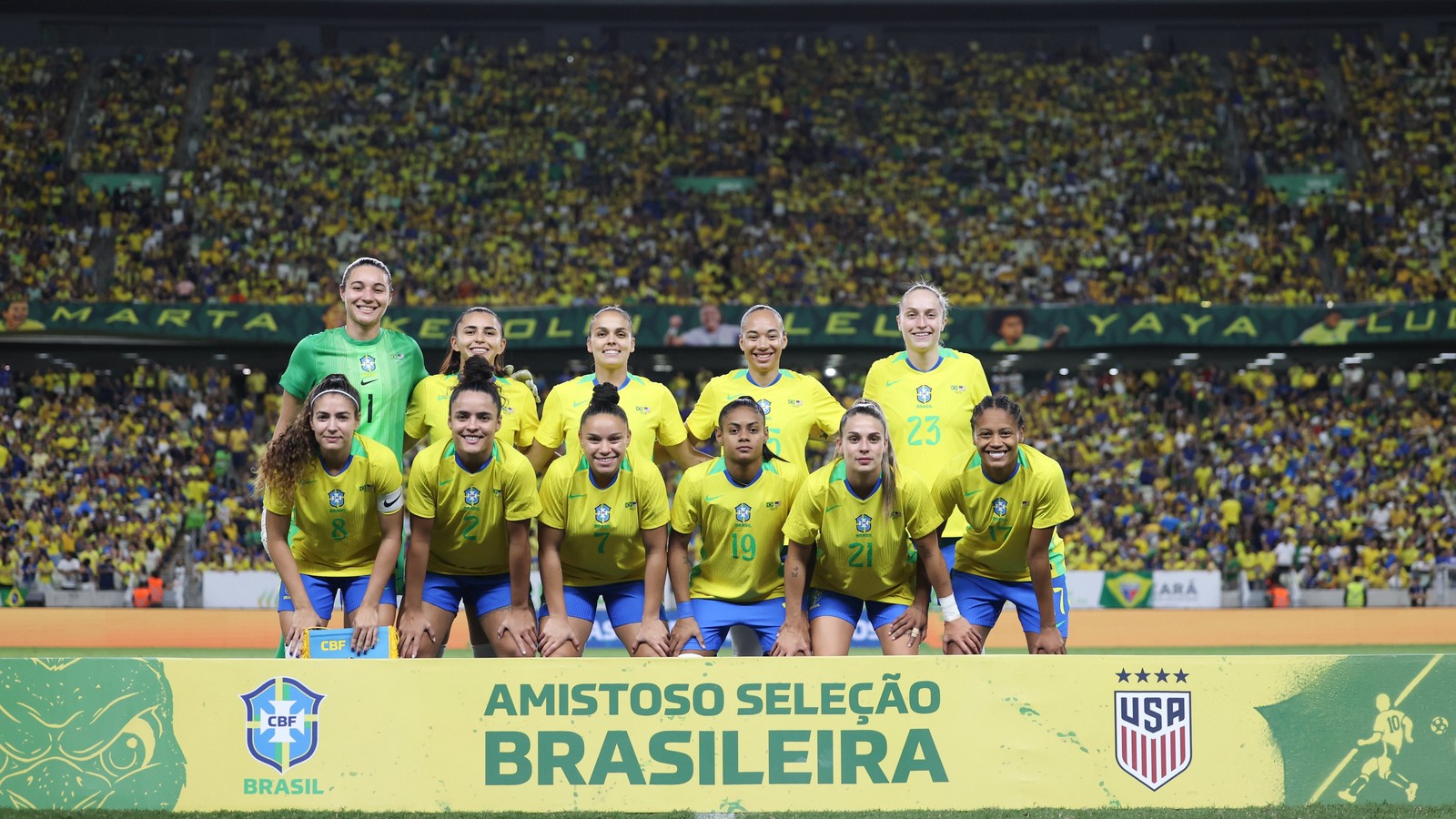 Seleção brasileira feminina posa em campo, estádio lotado. Faixa com "AMISTOSO SELEÇÃO BRASILEIRA", CBF e USA