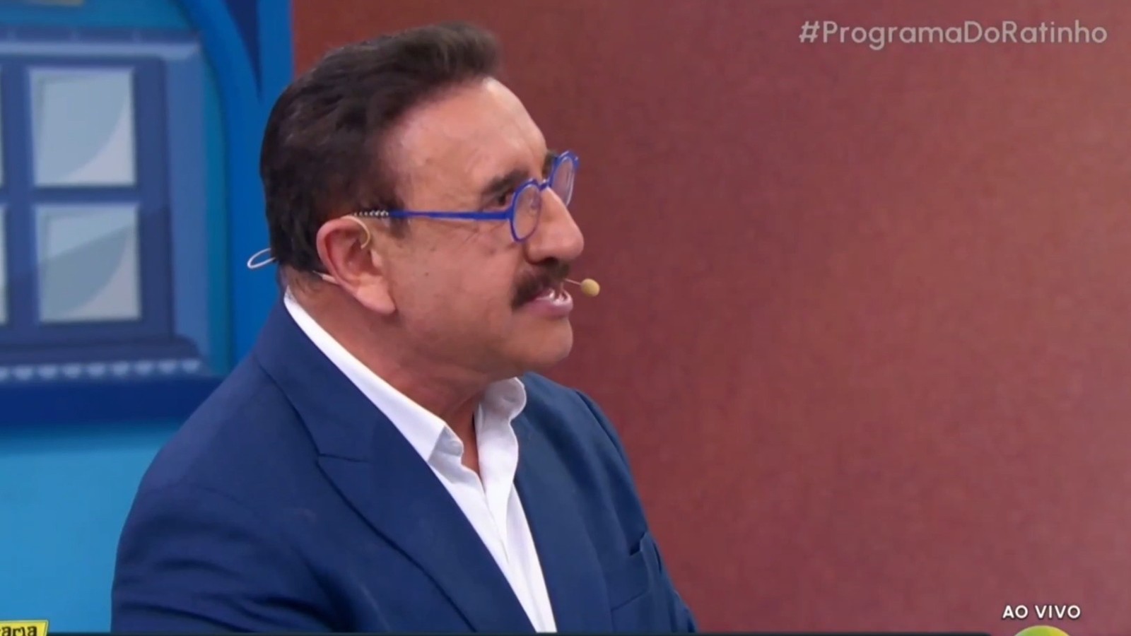 Ratinho de perfil, com bigode, óculos azuis, terno. Microfone no rosto. Textos #ProgramaDoRatinho, AO VIVO