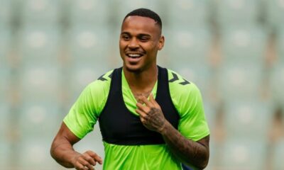 Homem sorridente com camisa verde e colete esportivo preto em campo de futebol