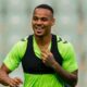Homem sorridente com camisa verde e colete esportivo preto em campo de futebol