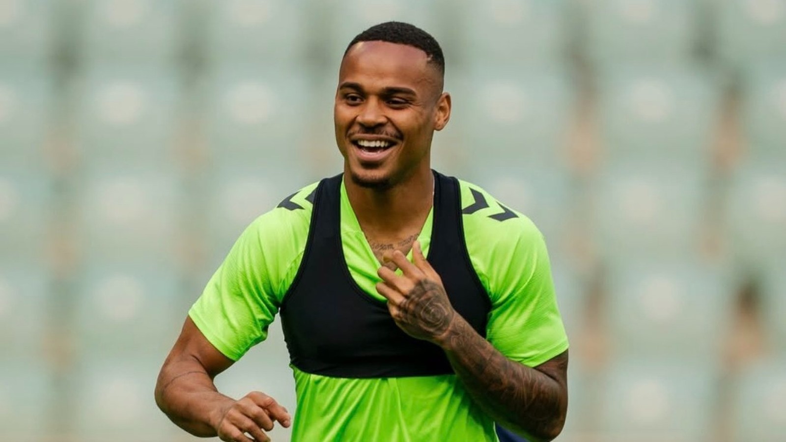 Homem sorridente com camisa verde e colete esportivo preto em campo de futebol