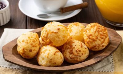 Pães de queijo redondos e dourados em travessa de madeira, com bebidas e geleia no fundo