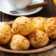 Pães de queijo redondos e dourados em travessa de madeira, com bebidas e geleia no fundo