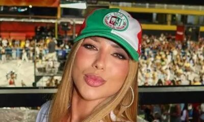 Mulher loira de boné verde, branco e vermelho, com maquiagem e bico, em evento.