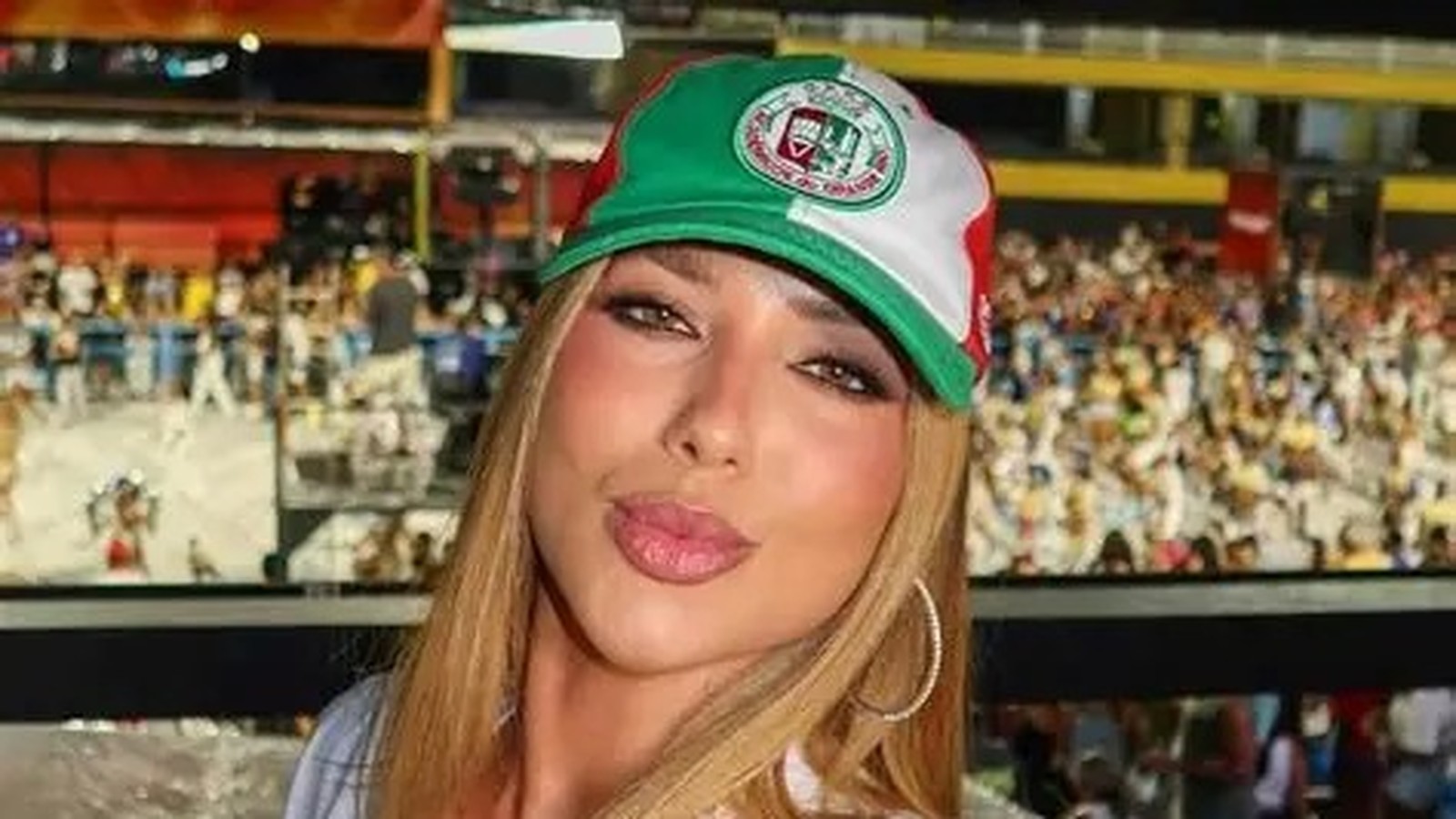 Mulher loira de boné verde, branco e vermelho, com maquiagem e bico, em evento.
