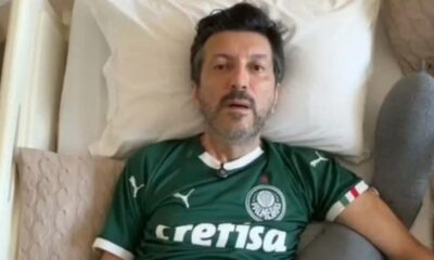 Homem deitado, cabelo escuro e barba, vestindo camisa verde do Palmeiras, cercado por travesseiros