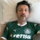 Homem deitado, cabelo escuro e barba, vestindo camisa verde do Palmeiras, cercado por travesseiros