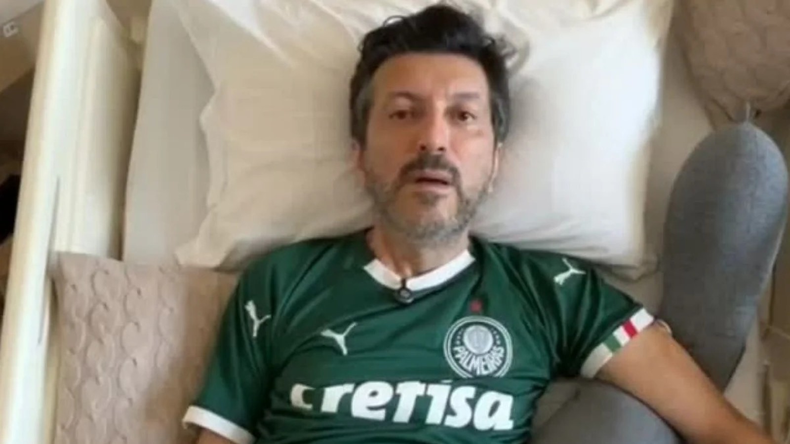 Homem deitado, cabelo escuro e barba, vestindo camisa verde do Palmeiras, cercado por travesseiros