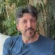 Homem de meia-idade com barba grisalha e camiseta azul em ambiente com plantas e janela